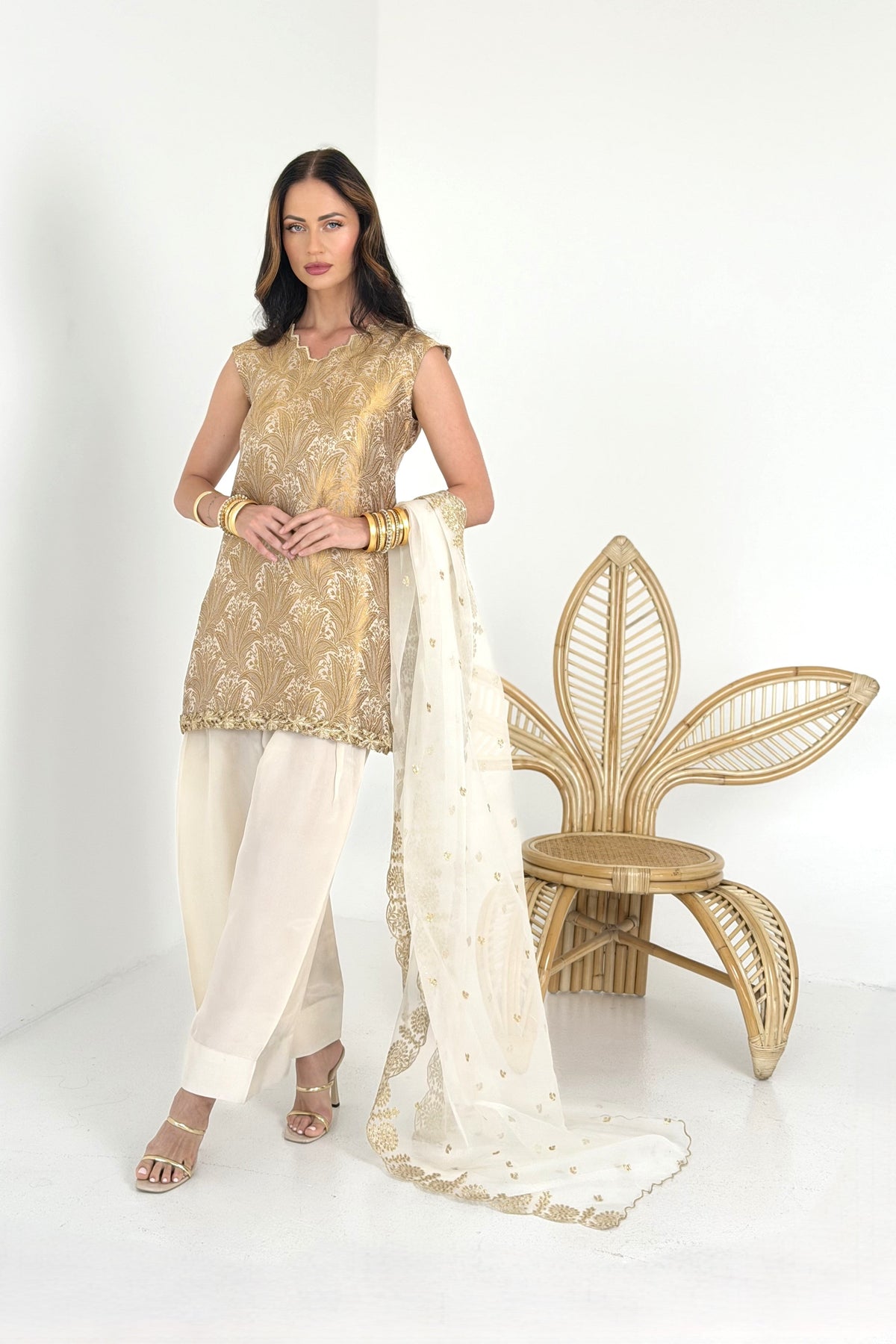 A woman models a traditional South Asian outfit consisting of a gold embroidered kurta, cream pants, and a white dupatta.