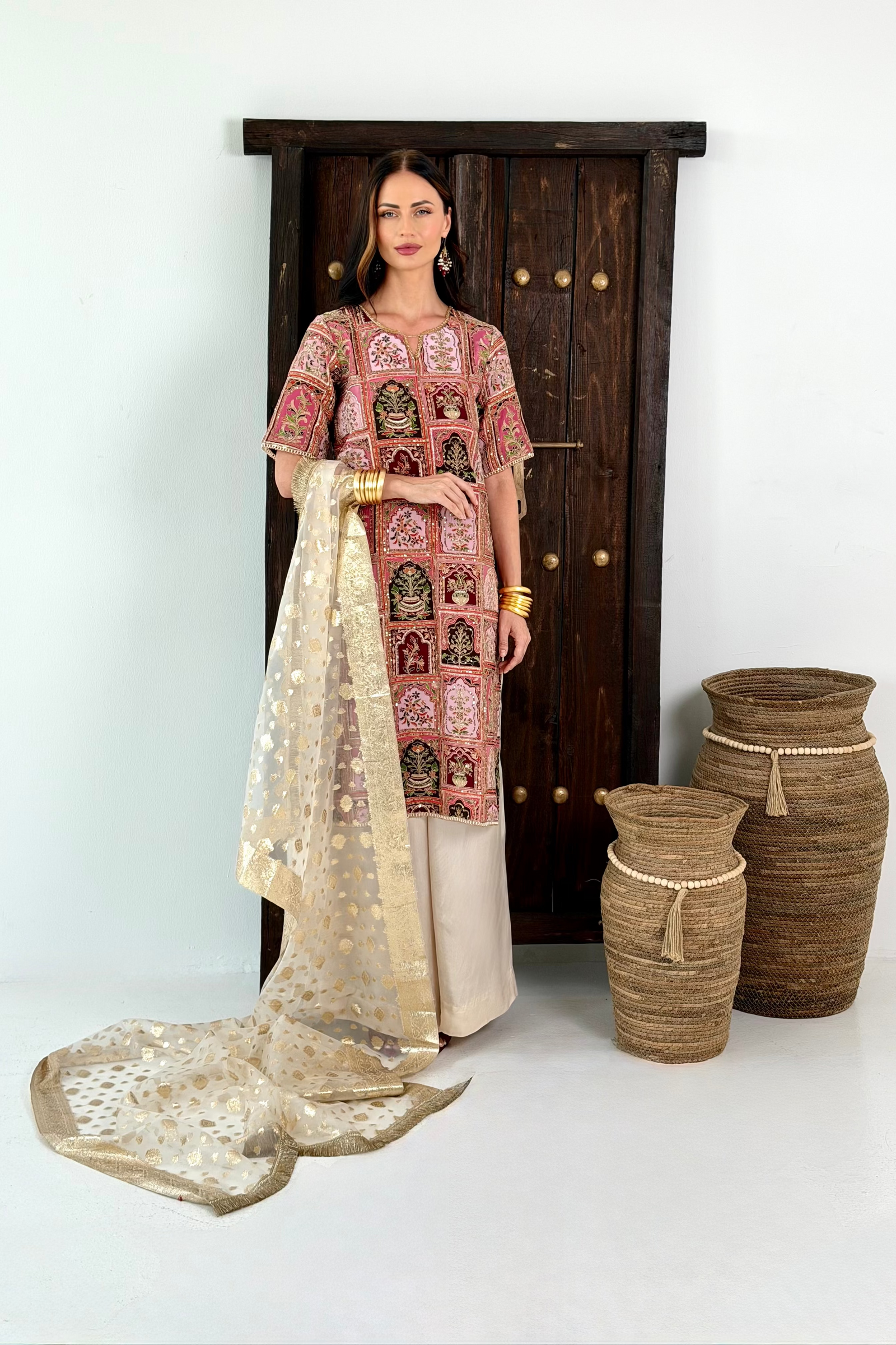 A woman models a traditional South Asian salwar kameez suit set consisting of a pink and beige patterned kurta, beige trousers, and a sheer beige dupatta with gold motifs, standing in front of a dark wooden door with decorative woven baskets.