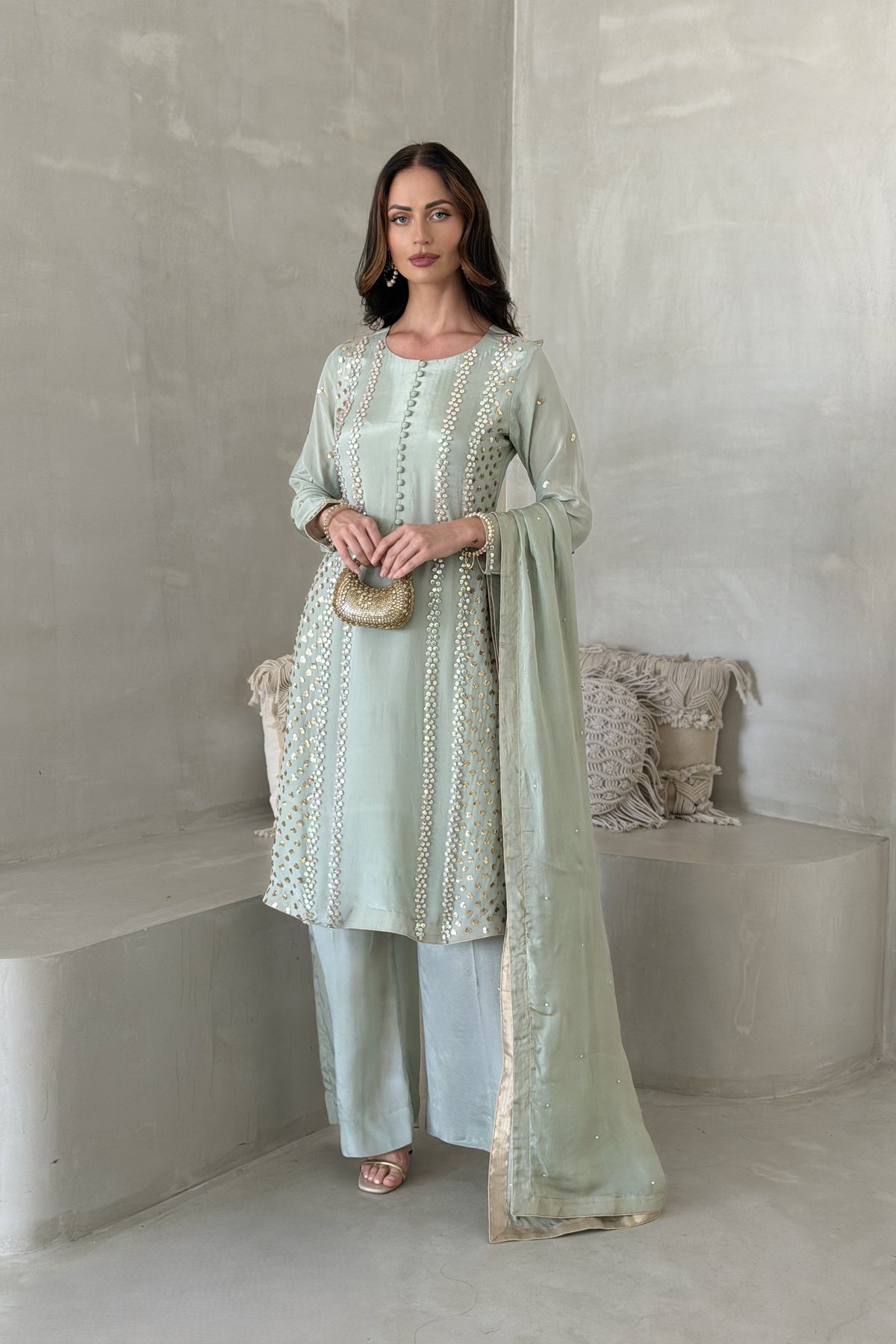 A woman models a light sage green traditional three-piece outfit with intricate gold embroidery, matching wide-leg trousers, and a coordinating dupatta.