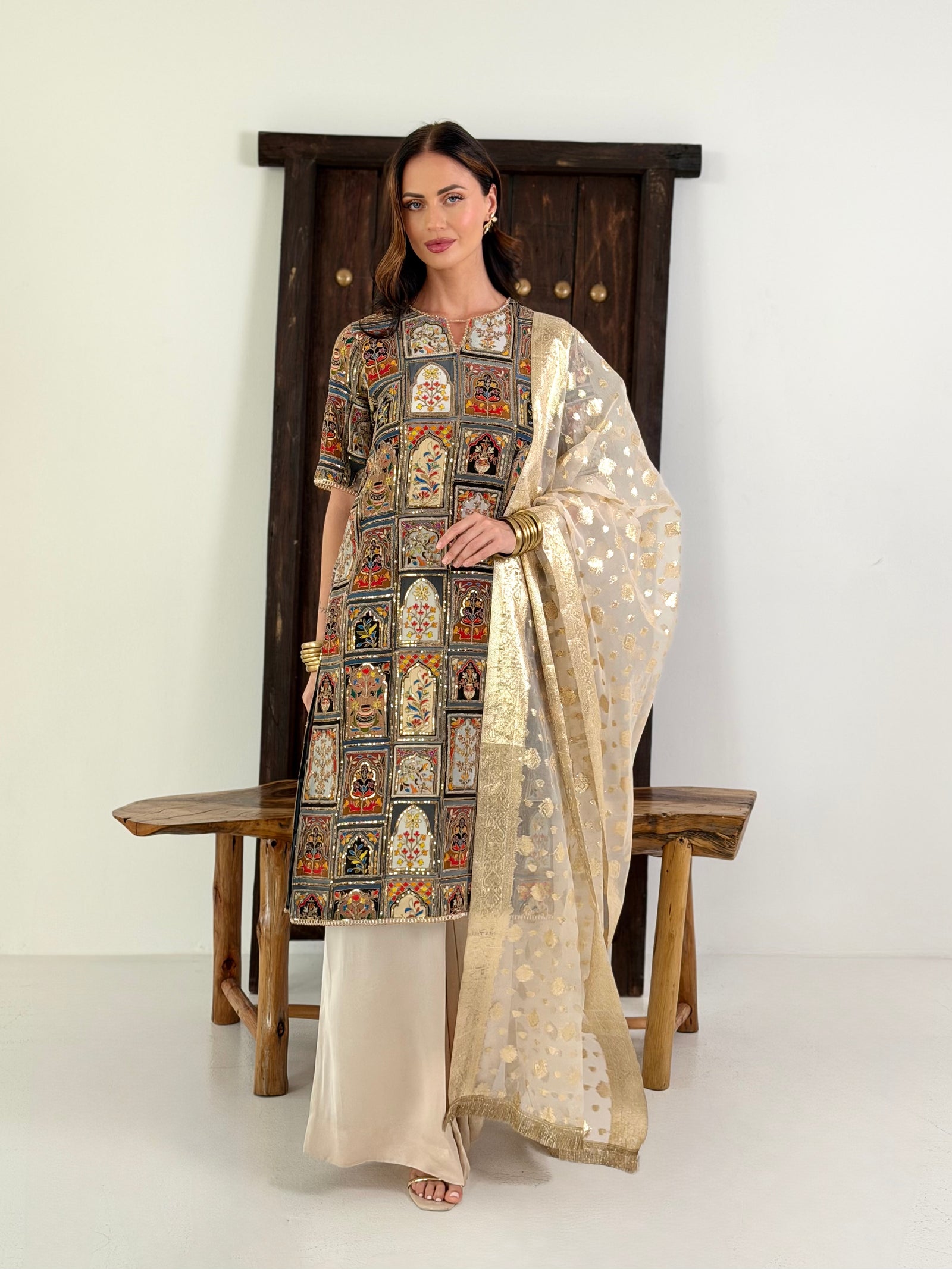 A woman models a traditional South Asian salwar kameez suit set consisting of a heavily embroidered kameez tunic, beige satin trousers, and a sheer beige dupatta with gold motifs, posed in front of a rustic wooden bench and door frame.